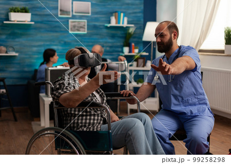 Elderly woman in wheelchair using virtual reality glasses in common room of nursing home, male nurse providing assistance and teaching how to use modern technology. Time for fun in care center. 99258298