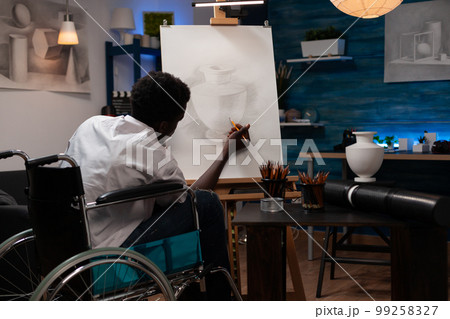 Young african american man with artistic abilities living with health condition making pencil sketch of vase at home art studio. Male wheelchair user spending time doing drawing task. Young african american man with artistic abilities living with health condition making pencil sketch of vase at home art studio. Male wheelchair user spending time doing drawing task. 99258327