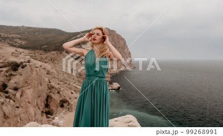 Redhead woman portrait. Curly redhead young caucasian woman with freckles looking at camera and smiling. Close up portrait cute woman in a mint long dress posing on a volcanic rock high above the sea 99268991