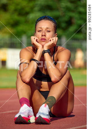 Fit woman in sportswear resting after training or running, sitting on a rubber treadmill, taking a break during training, outdoors. 99285484