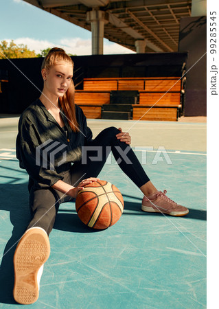 Girl basketball player smiles and sits on the sports field with a basketball and looks at the camera. Sport, fitness, active lifestyle. 99285545