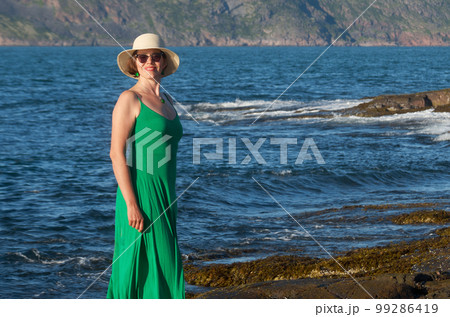 Woman tourist in a straw hat and a green dress on the seashore on a bright sunny day 99286419