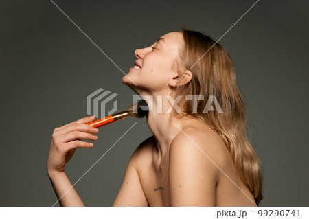 Profile view portrait of young beautiful girl with bare shoulders powdering with makeup brush. Concept of beauty, natural make-up, skin care, healthy look 99290741