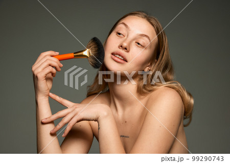 Half-length portrait of young beautiful girl with bare shoulders powdering with makeup brush. Concept of beauty, natural make-up, skin care, healthy look 99290743