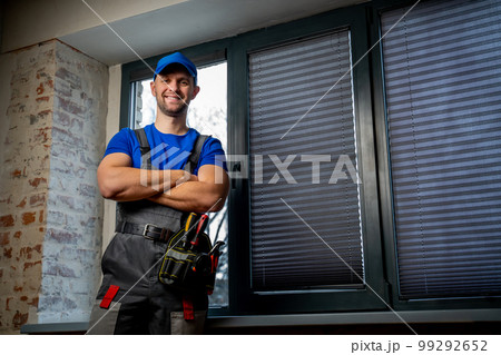 Maintenance worker in uniform, blue T-shirt and cap, crossing his arms. A handyman wearing a tool belt against a black window background Maintenance worker in uniform, blue T-shirt and cap, crossing his arms. A handyman wearing a tool belt against a black window background 99292652