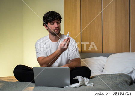 Man who feels sick, sitting at home on a bed, drips nasal drops into a stuffy nose. A man sprays cold medicine for a runny nose and listens to the doctor using a laptop Man who feels sick, sitting at home on a bed, drips nasal drops into a stuffy nose. A man sprays cold medicine for a runny nose and listens to the doctor using a laptop 99292718