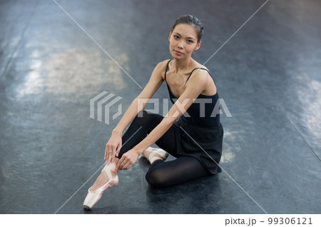 An Asian woman dressed in a black bodysuit, leggings and pointe shoes sits on the floor. 99306121