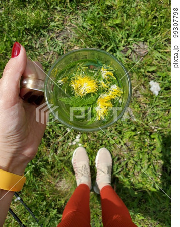 A woman holding a glass mug of herbal tea with dandelion on the grass on a sunny day 99307798