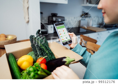 Online home food delivery. Focus on active app with order list on the phone screen. Woman checking her order. Cardboard box with fresh vegetables standing on the kitchen table. Local farmer food. 99312176