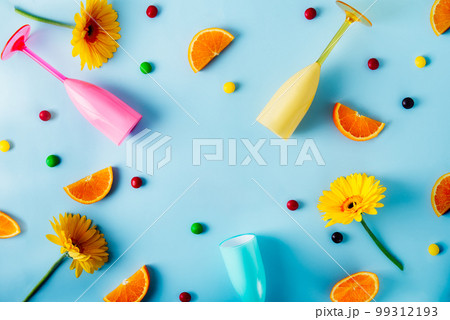 Top view background with bright plastic cocktail glasses, multicolor candy drops, orange slices and yellow gerbera flowers on the light blue background. Summer party mood. Flatlay, copy space. 99312193