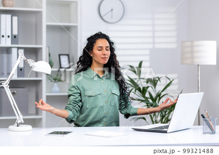 Young beautiful hispanic woman relaxing in home office, successful businesswoman meditating in lotus position sitting at workplace with laptop, freelancer in green shirt in living room. Young beautiful hispanic woman relaxing in home office, successful businesswoman meditating in lotus position sitting at workplace with laptop, freelancer in green shirt in living room. 99321488
