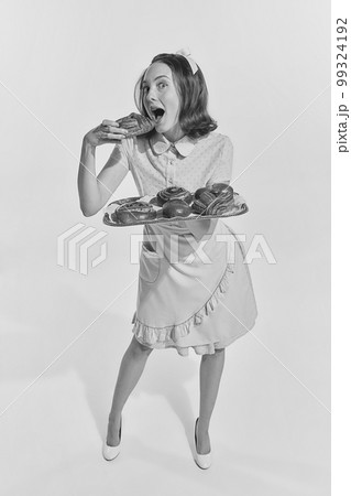 Black and white portrait of young redhead woman in cute pink dress biting freshly baked buns. Vintage, retro style 99324192
