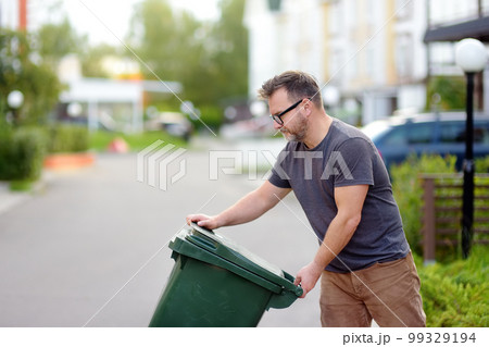 Mature man pulling out a large green plastic garbage container in front of the townhouse to the roadway of the street. Garbage collection and recycling trash Mature man pulling out a large green plastic garbage container in front of the townhouse to the roadway of the street. Garbage collection and recycling trash 99329194