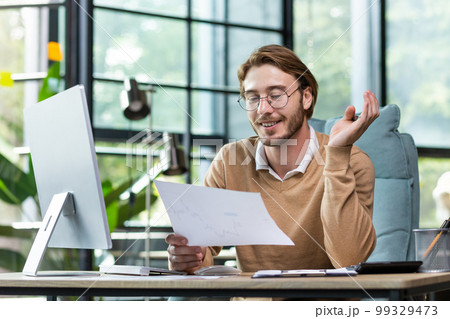 A young male student sits in the office and holds documents in his hands, studies, works at the computer, smiles. A young male student sits in the office and holds documents in his hands, studies, works at the computer, smiles. 99329473