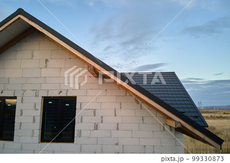 Unfinished house with aerated lightweight concrete walls and wooden roof frame covered with metallic tiles under construction Unfinished house with aerated lightweight concrete walls and wooden roof frame covered with metallic tiles under construction 99330873