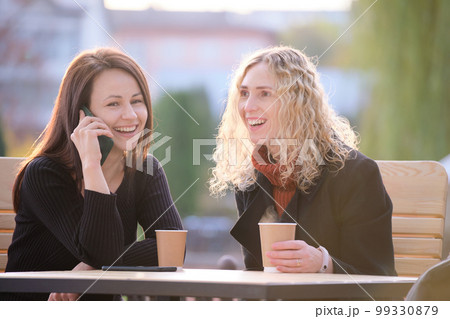 Two happy women friends having fun time together at street cafe laughing and talking on cellphone Two happy women friends having fun time together at street cafe laughing and talking on cellphone 99330879