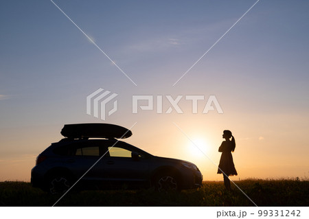 Silhouette of female driver standing near her car on grassy field enjoying view of bright sunset. Young woman relaxing during road trip beside SUV vehicle. 99331242