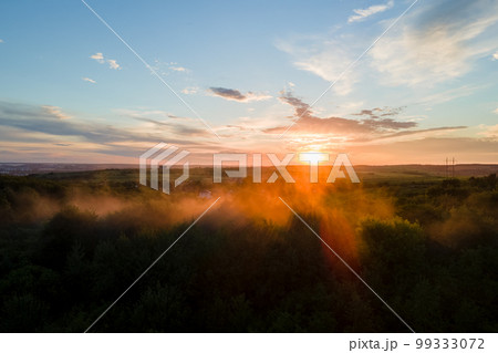 Aerial view of light fog covering dark forest trees at warm sunset. 99333072
