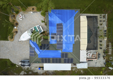 Aerial view of damaged in hurricane Ian house roof covered with blue protective tarp against rain water leaking until replacement of asphalt shingles Aerial view of damaged in hurricane Ian house roof covered with blue protective tarp against rain water leaking until replacement of asphalt shingles 99333376