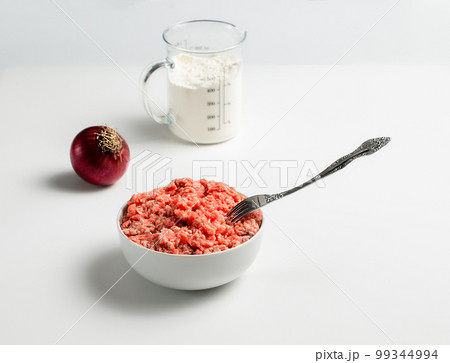Plate with fresh ground beef and fork, red onion, measuring cup with flour on a white background, space for text, stock photo Plate with fresh ground beef and fork, red onion, measuring cup with flour on a white background, space for text, stock photo 99344994
