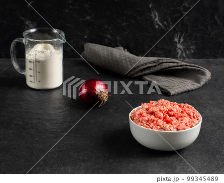 Plate with fresh ground beef, red onion, measuring cup with flour on a gray black background, space for text, stock photo Plate with fresh ground beef, red onion, measuring cup with flour on a gray black background, space for text, stock photo 99345489