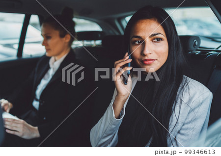Young business woman witting in the back of a car with a colleague, talking on the phone and looking out the window 99346613