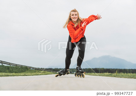 View from the front young woman in red jacket skating fast towards the camera with a big smile on her face  99347727