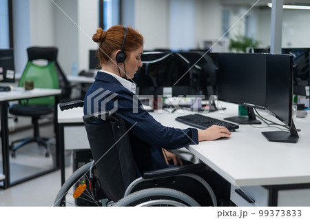 Red-haired caucasian woman in a wheelchair talking on a headset. Female call center worker at her desk. 99373833