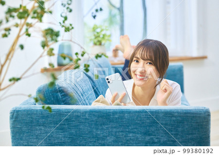 ソファーでうつ伏せになりながらスマホを操作する若い女性 ソファーでうつ伏せになりながらスマホを操作する若い女性 99380192