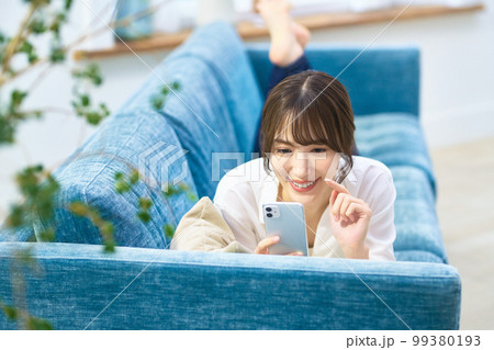 ソファーでうつ伏せになりながらスマホを操作する若い女性 ソファーでうつ伏せになりながらスマホを操作する若い女性 99380193