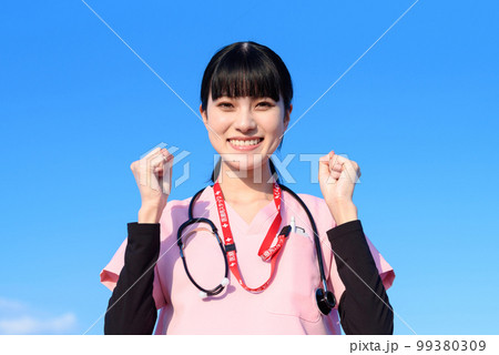 青空を背景にして立つスクラブを着た女性看護師　医療従事者　医師　ガッツポーズ　トリミングアップ 99380309
