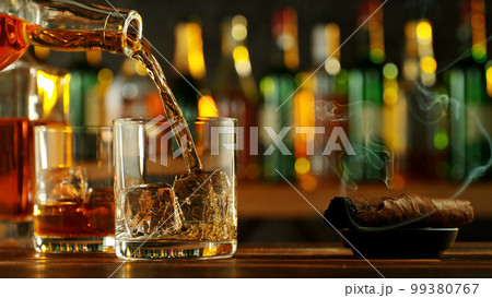 Detail of pouring whiskey into glass with ice cubes. 99380767