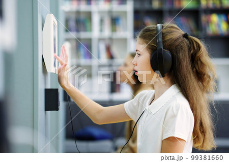 School girl with long hair wearing white t-shirt studying in class. Side view student with headphones using device with touchscreen on wall, another child on blurred background. Concept of education School girl with long hair wearing white t-shirt studying in class. Side view student with headphones using device with touchscreen on wall, another child on blurred background. Concept of education 99381660