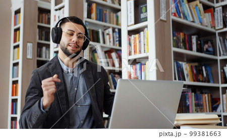 Bearded man with eyeglasses wearing headphones working on laptop in library, starting singing and dancing when hearing popular music. Student taking break in studying and having fun 99381663