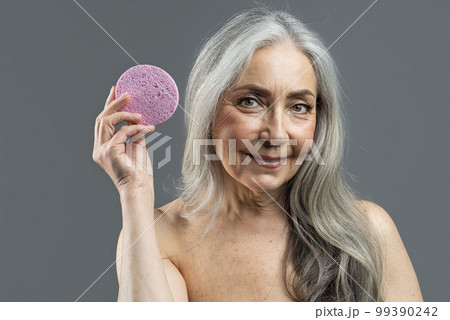Smiling retired european naked female with gray hair shows massage sponge for face 99390242