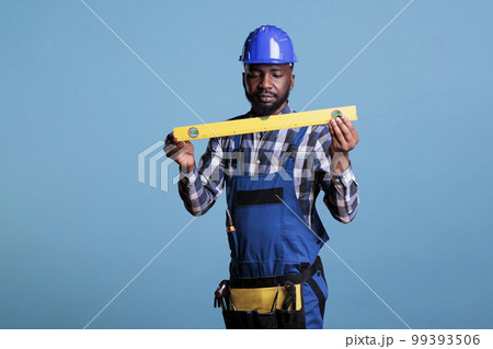 Concentrated african american construction worker in uniform holding leveler on blue background. Experienced builder carrying work tools in belt and hard hat, studio shot. 99393506