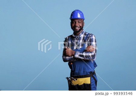 African american construction worker with happy face wearing a helmet smiling while looking at camera. Confident builder posing with arms crossed holding wrench against blue background. 99393546