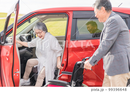 自動車から降りて車椅子に乗る高齢者女性と介助する男性（移乗） 99396648