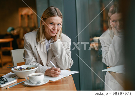 Smiling businesswoman having lunch and making notes while working in cafe Smiling businesswoman having lunch and making notes while working in cafe 99397973