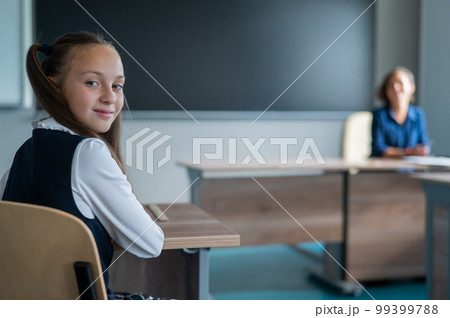 Caucasian girl and her teacher in the classroom. Schoolgirl turns around and looks at the camera. Caucasian girl and her teacher in the classroom. Schoolgirl turns around and looks at the camera. 99399788