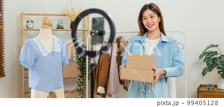 Female online seller is using smartphone to broadcast live on Vlog for introduce fashions and holding 99405128