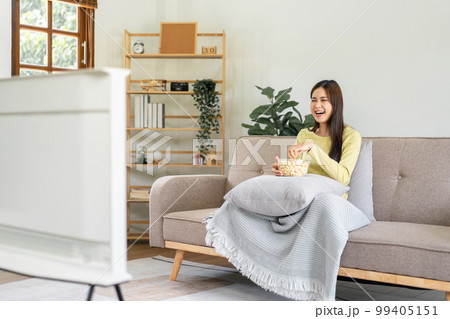 Young asian woman putting a bowl of popcorn on pillow and eating popcorn while sitting on the big comfortable Young asian woman putting a bowl of popcorn on pillow and eating popcorn while sitting on the big comfortable 99405151