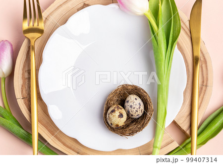 White figured wavy plate, two quail eggs in nest, golden cutlery, pink tulips on round wooden stand. Easter table setting. Festive mock up close up. Top view. Copy space. White figured wavy plate, two quail eggs in nest, golden cutlery, pink tulips on round wooden stand. Easter table setting. Festive mock up close up. Top view. Copy space. 99407314