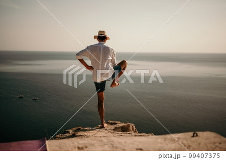 A digital nomad, a man in a hat, a businessman does yoga on the rocks by the sea at sunset time, does a business operation online from a distance. Remote work on vacation. 99407375