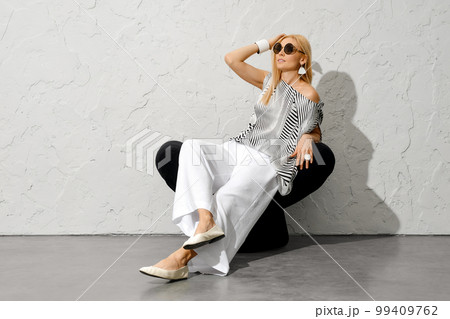 Charming woman in sunglasses, striped blouse and wide trousers sitting on armchair Charming woman in sunglasses, striped blouse and wide trousers sitting on armchair 99409762