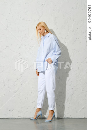 Pensive blonde woman in blue shirt, white jeans and stilettos looking down 99409765