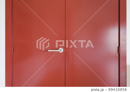 Closeup of a wide front double-leaf wooden door covered with red paint. Door handle made of durable metal. The door is held on door hinges. Sunlight sets off the depth of color. Closeup of a wide front double-leaf wooden door covered with red paint. Door handle made of durable metal. The door is held on door hinges. Sunlight sets off the depth of color. 99410956