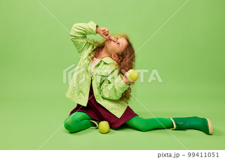 Having fun. Little cute girl, child with curly hair posing with apples over green studio background. Concept of childhood, emotions, fun Having fun. Little cute girl, child with curly hair posing with apples over green studio background. Concept of childhood, emotions, fun 99411051