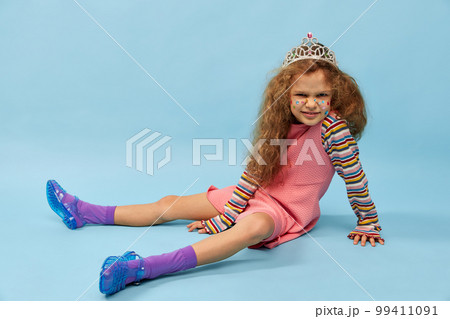 Serious face. Little cute girl, child with curly hair posing in pink dress and crown over blue studio background. Concept of childhood, emotions, fun Serious face. Little cute girl, child with curly hair posing in pink dress and crown over blue studio background. Concept of childhood, emotions, fun 99411091