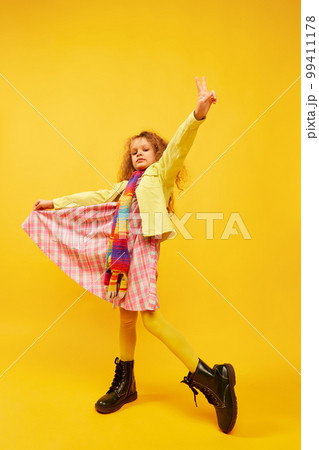 Little cute girl, child with curly hair posing in bright clothes, scarf and boots over yellow studio background. Concept of childhood, emotions, fun Little cute girl, child with curly hair posing in bright clothes, scarf and boots over yellow studio background. Concept of childhood, emotions, fun 99411178